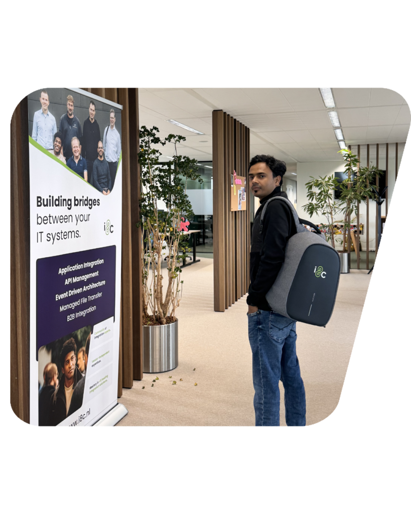 Picture of Sagar, solution architect at i8c NL, next to a banner of i8c NL, in our Utrecht office with a backpack of the company